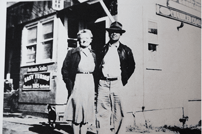 Two men in front of a building - one in suit and hat, one in overalls and cap